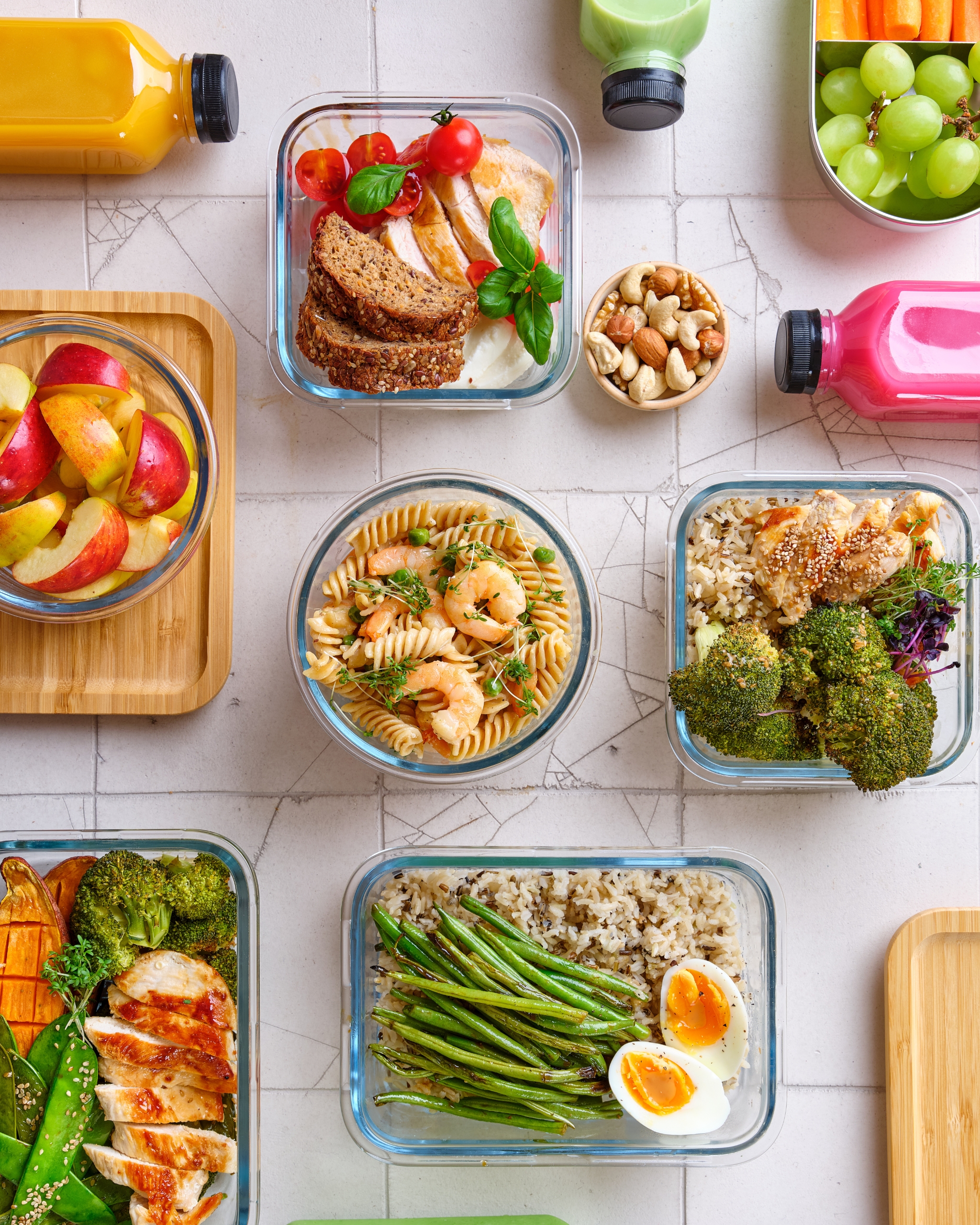 An overhead view of various meal planning containers filled with healthy foods. The containers include pasta salad, sandwiches, fruits, vegetables, nuts, and proteins like eggs and chicken. Colorful drink bottles are also visible, suggesting a well-planned and balanced meal prep setup.