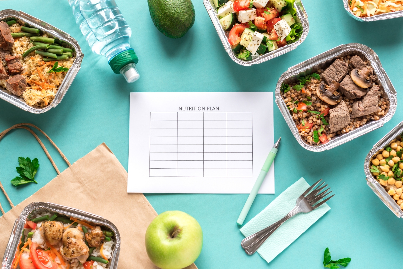 Overhead view of a meal planning setup on a teal background. Various meal prep containers with healthy foods, a nutrition plan sheet, water bottle, apple, utensils, and a paper bag are arranged around a central nutrition plan template.