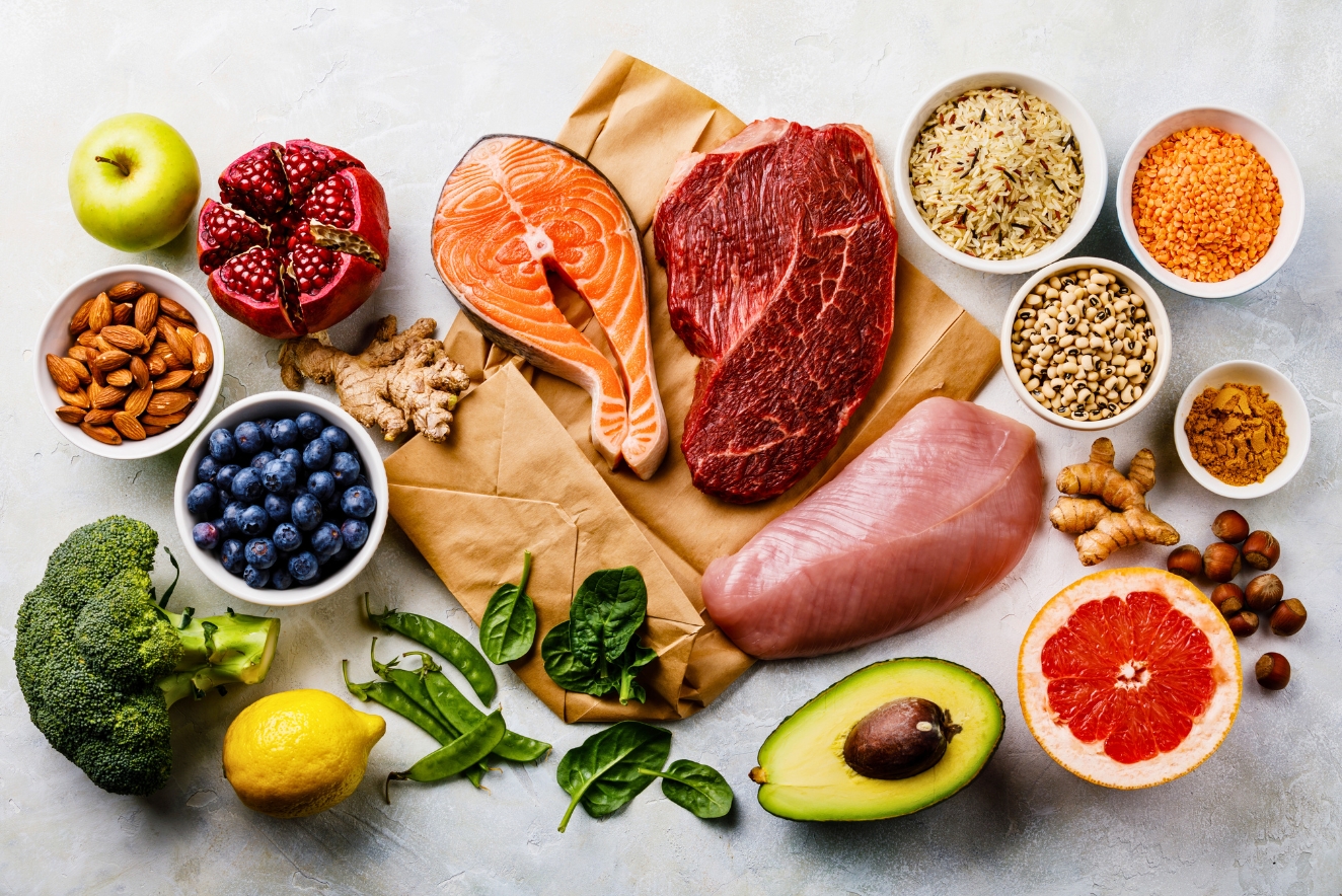 A variety of nutrient-rich foods displayed on a light background. The image includes meats (beef, salmon, chicken), fruits (apple, pomegranate, blueberries, grapefruit, lemon), vegetables (broccoli, avocado, spinach), nuts, grains, and spices. The foods represent a balanced diet with diverse macronutrients and micronutrients.