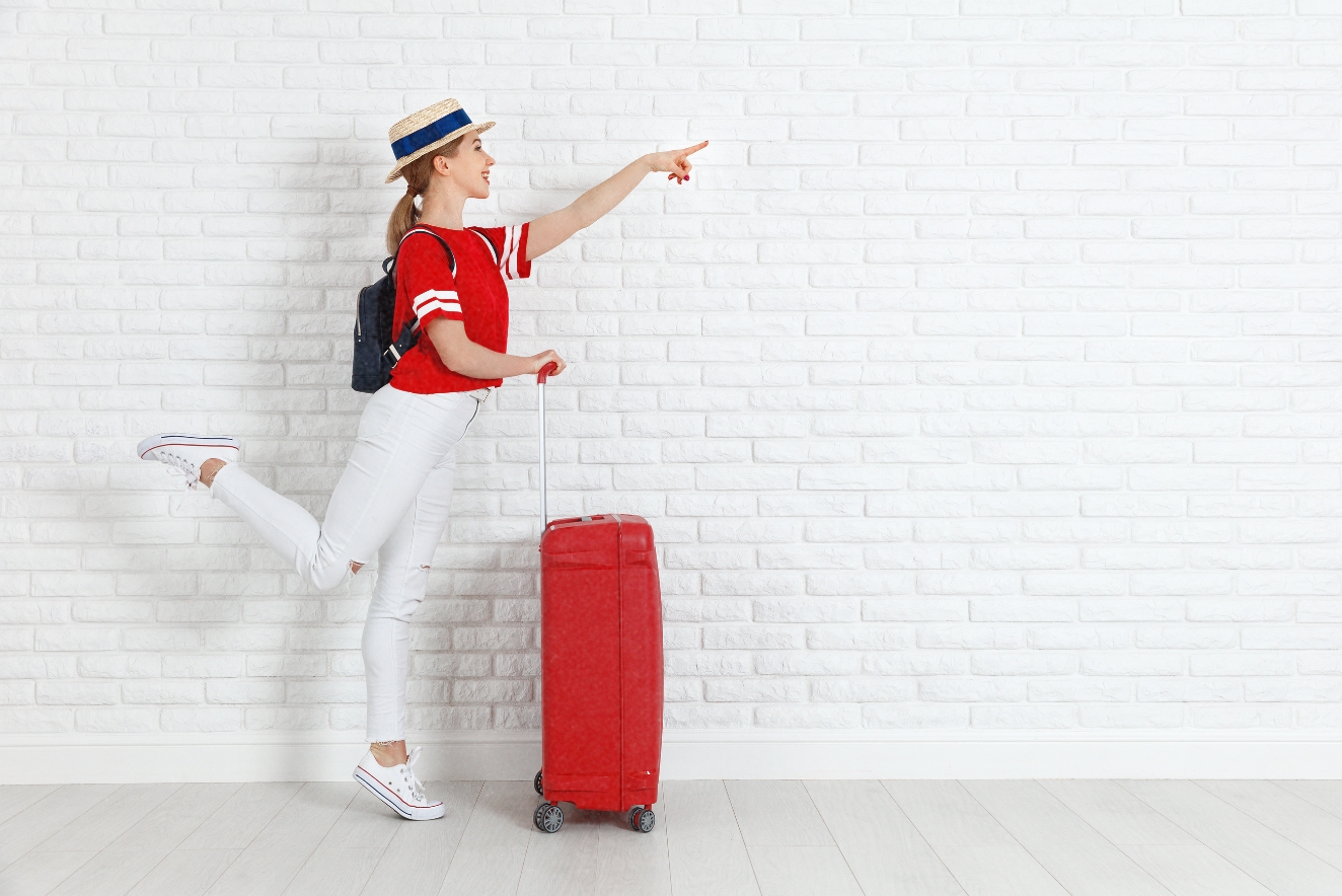 Budget travel excitement: Young woman in red shirt and hat gesturing enthusiastically next to a red suitcase, symbolizing affordable adventure preparation.