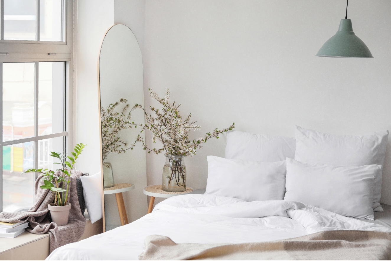 Home organization exemplified in a serene bedroom with a neatly made white bed, minimalist decor, and organized storage solutions including a mirror and plants.