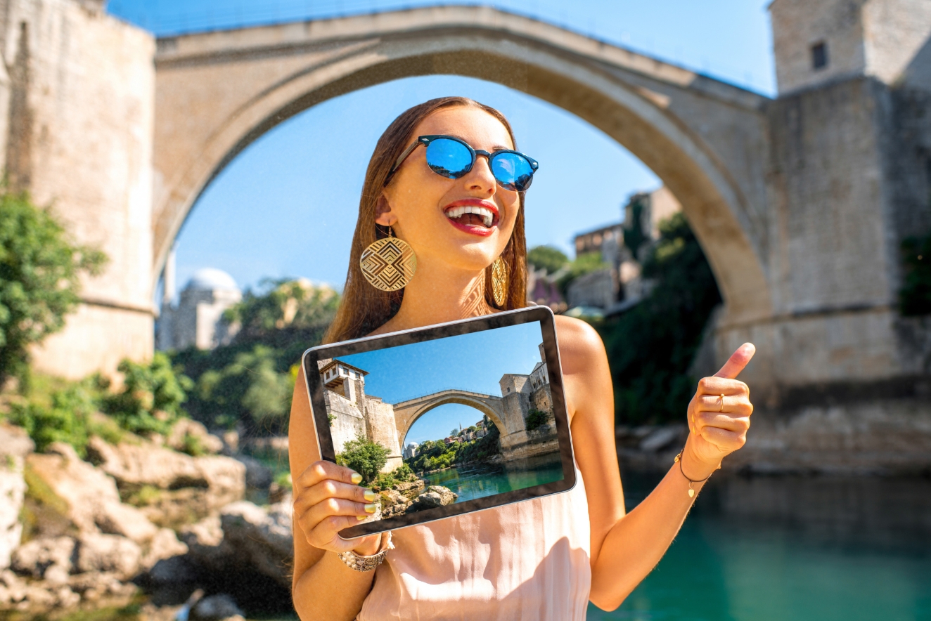 A smiling young woman tourist with sunglasses holds a tablet displaying a photo of the Stari Most bridge in Mostar, Bosnia and Herzegovina. She gives a thumbs up, demonstrating the use of digital apps for sustainable tourism and enhancing travel experiences. This image effectively illustrates the concept of using technology and apps to support sustainable tourism. The tourist is using a tablet to capture or view information about the historic Stari Most bridge, which suggests the use of digital guides or educational apps that can enhance a traveler's understanding of cultural sites without the need for printed materials. This aligns with the sustainable tourism practice of reducing paper waste and promoting digital solutions for travel information and experiences.