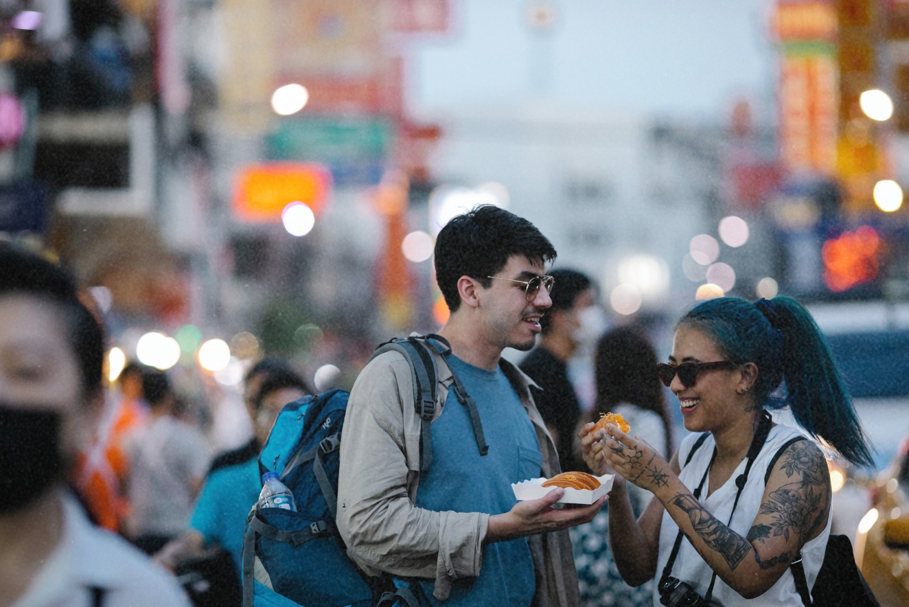  Two young travelers with backpacks enjoying street food in a bustling urban area, demonstrating sustainable eating habits by consuming local cuisine and reducing food waste while traveling.