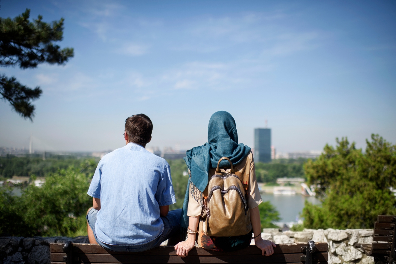 A couple, one wearing a hijab, sits on a stone wall overlooking a city skyline, exemplifying cultural respect and community engagement in sustainable tourism. 