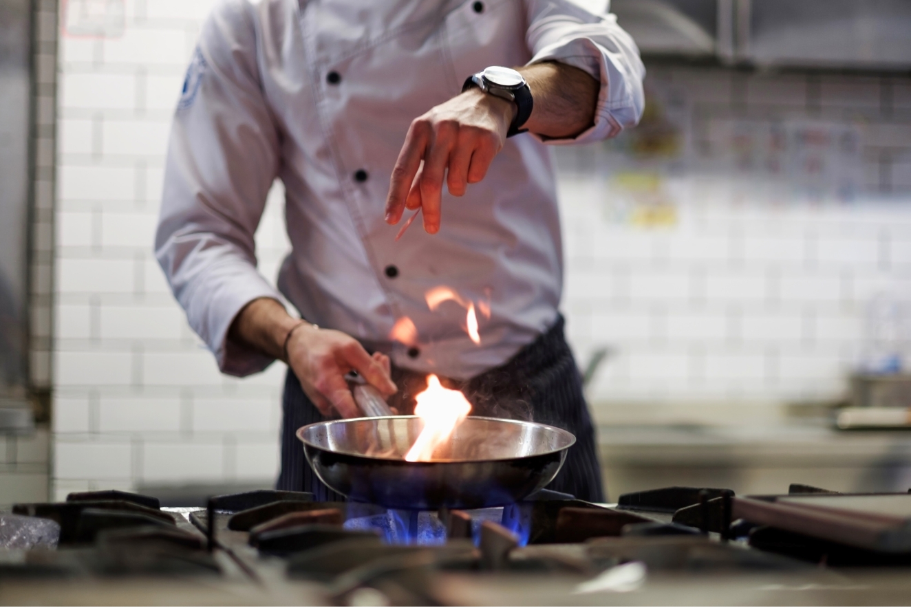 Chef flambéing a dish in a professional kitchen, demonstrating advanced cooking techniques with flames rising from a pan on a gas stove.