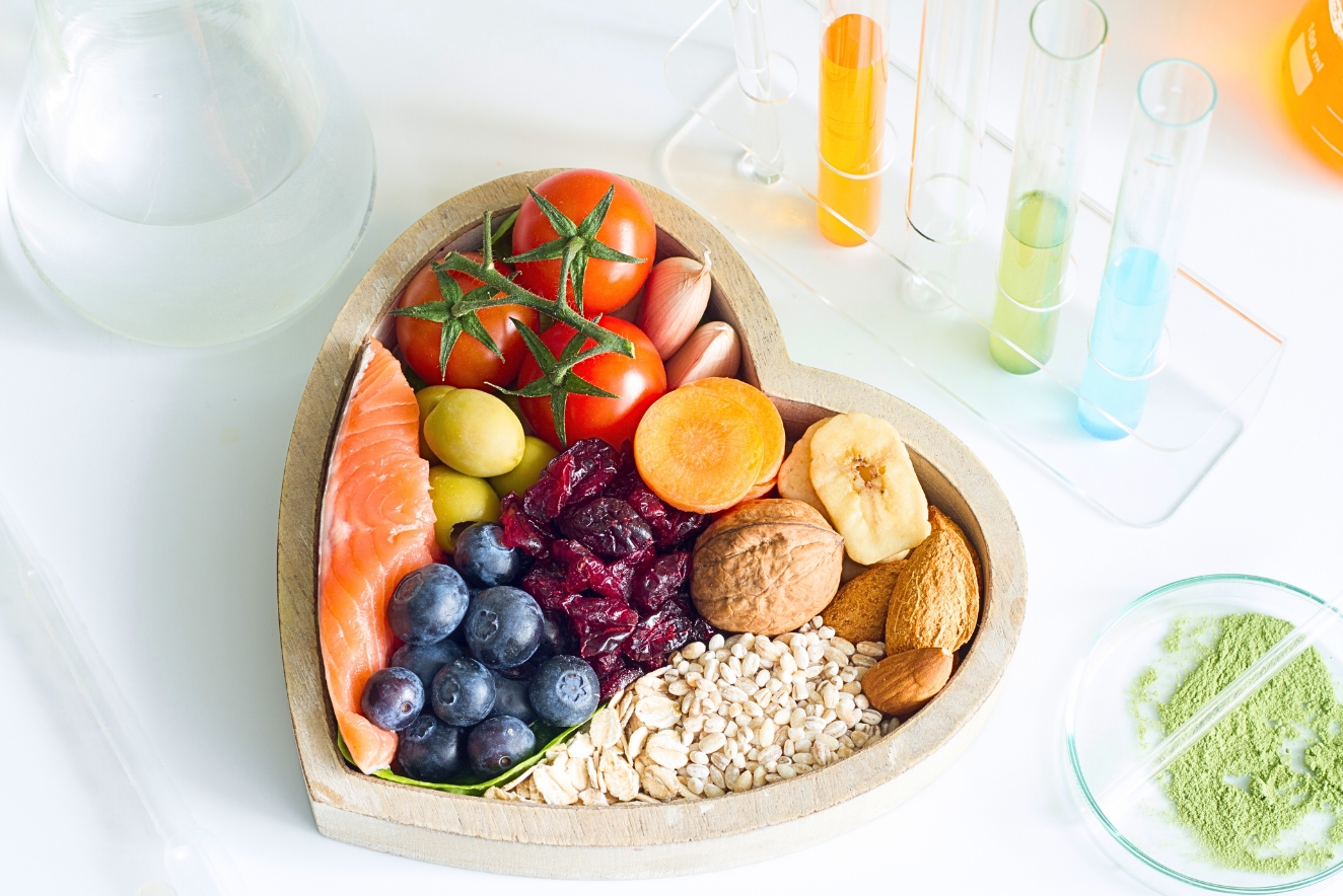 A heart-shaped wooden platter filled with a variety of healthy foods including salmon, tomatoes, blueberries, nuts, oats, dried fruit, and vegetables. Colorful liquid-filled test tubes and a dish of green powder are visible in the background, suggesting scientific analysis of nutrients.