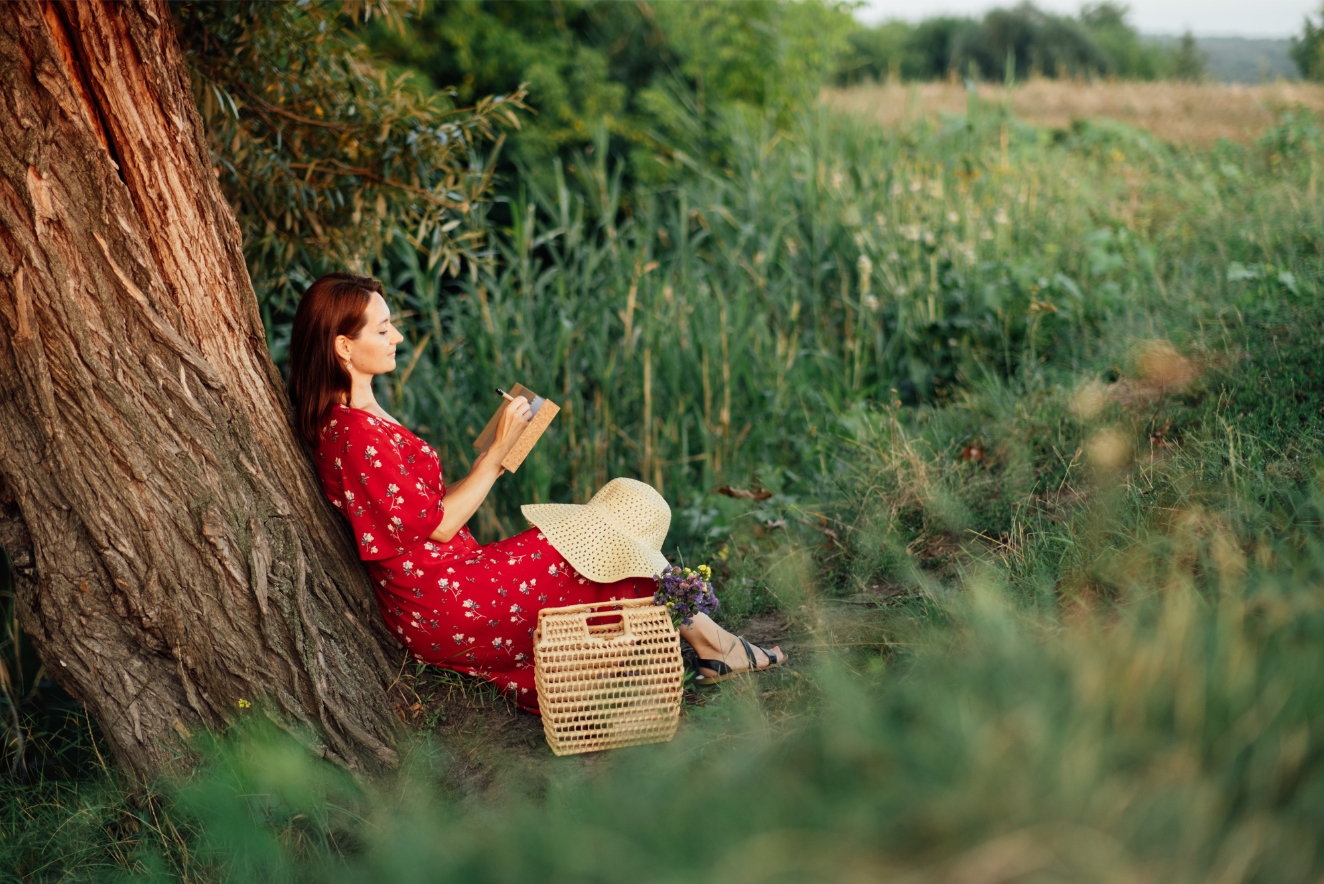 Woman practicing holistic health through nature connection and sketching outdoors, leaning against tree in serene field