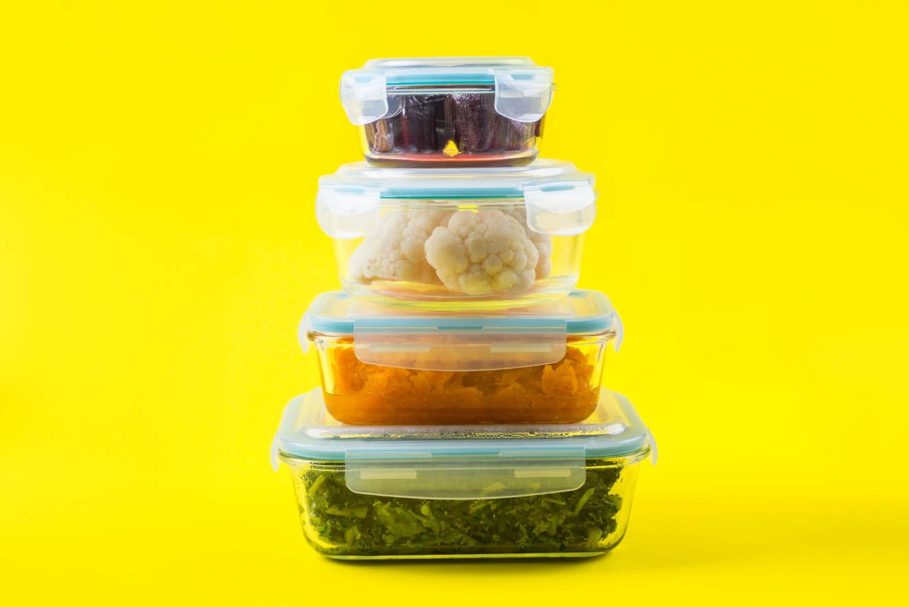 A stack of four transparent food storage containers with snap-on lids, filled with different foods including leafy greens, orange vegetables, cauliflower, and dark berries, arranged from largest at the bottom to smallest on top. The containers are set against a bright yellow background.