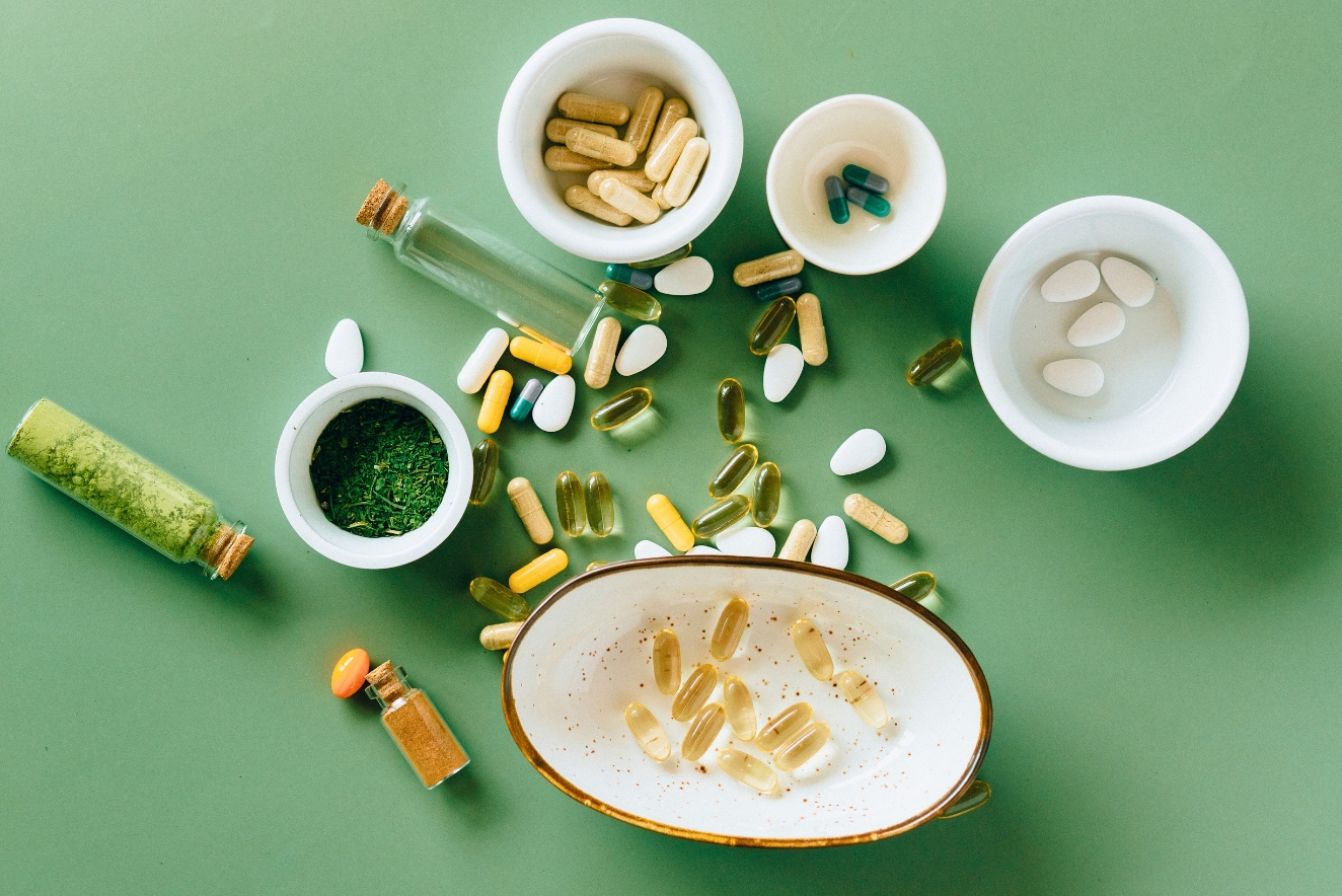  An assortment of dietary supplements and vitamins displayed on a green background. Various pills, capsules, and powders are scattered across the surface and contained in small white bowls. Two small glass bottles with liquid supplements are also visible.