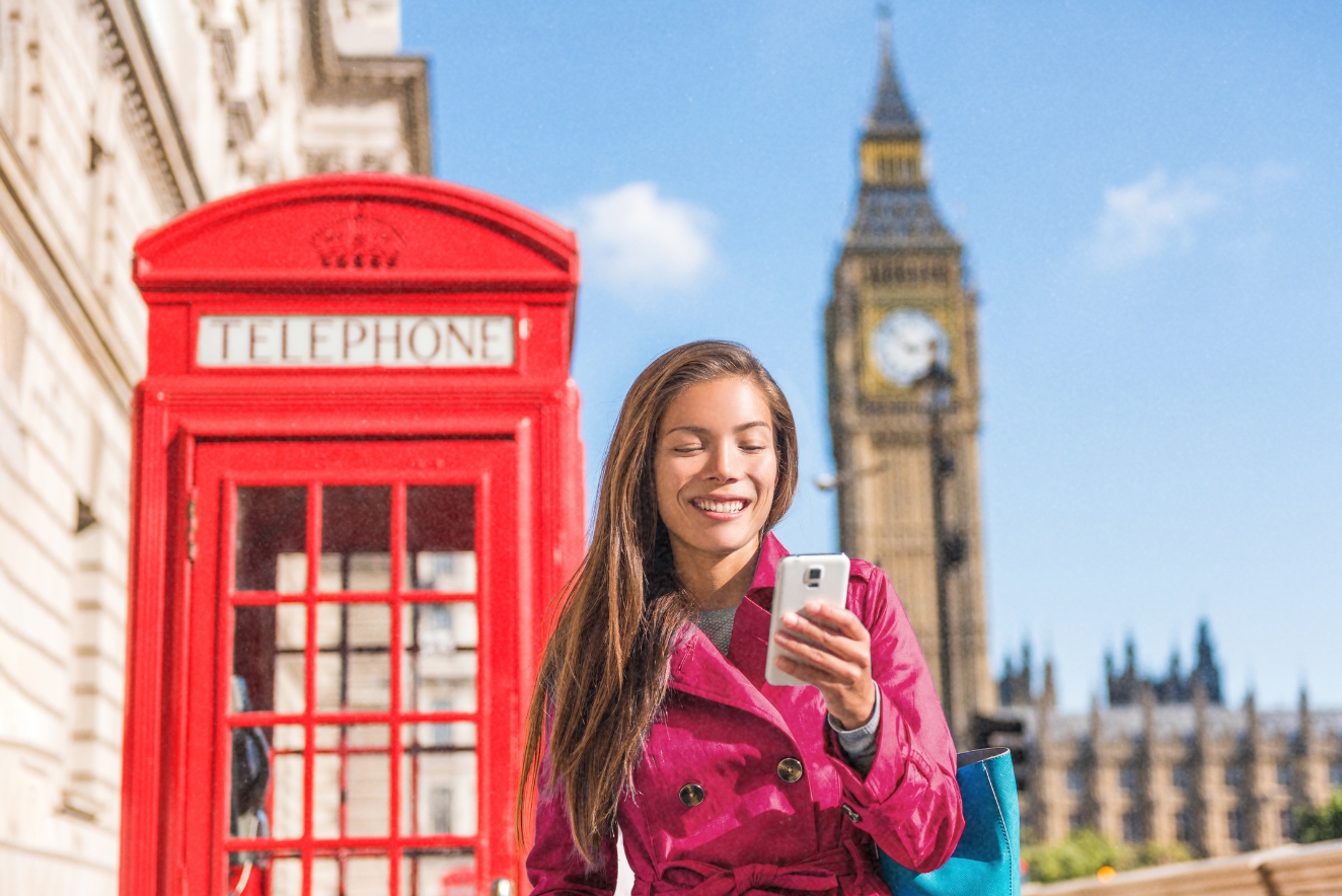 Budget travel tech: Tourist using smartphone app near London's iconic red phone booth and Big Ben, showcasing digital tools for affordable exploration.