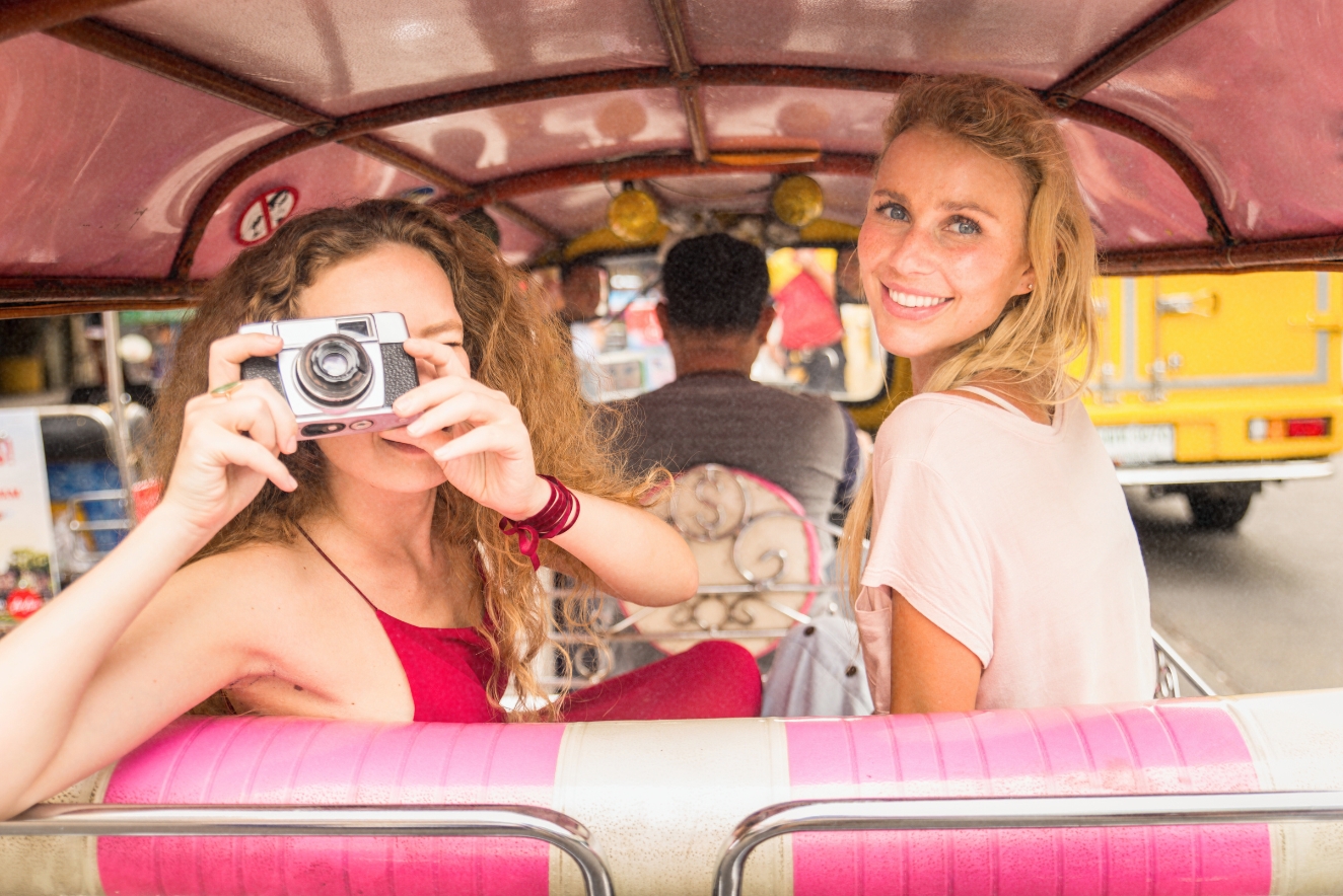 Budget travel adventure: Tourists using local transportation, with one woman taking a photo and another smiling inside a colorful tuk-tuk or auto-rickshaw.