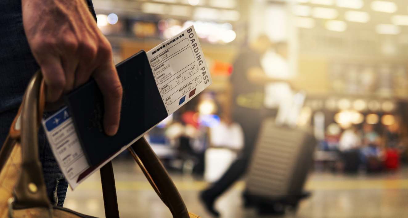 A close-up image of a hand holding a boarding pass against a blurred airport terminal background. The person is grasping the paper ticket while standing in what appears to be a busy departure area with other travelers and seating visible but out of focus in the background. The warm lighting creates an atmospheric travel scene that captures the moment before boarding a flight.