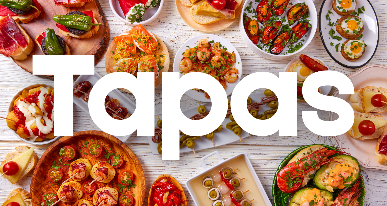 This image shows a variety of colorful Spanish tapas dishes arranged on a wooden table. The word "Tapas" is overlaid in large white text across the center of the image. The assortment includes what appears to be seafood, meats, and vegetable dishes in different serving plates and bowls, showcasing the diversity of Spanish small plates cuisine. The vibrant colors of the various dishes create an appetizing display of Mediterranean food.